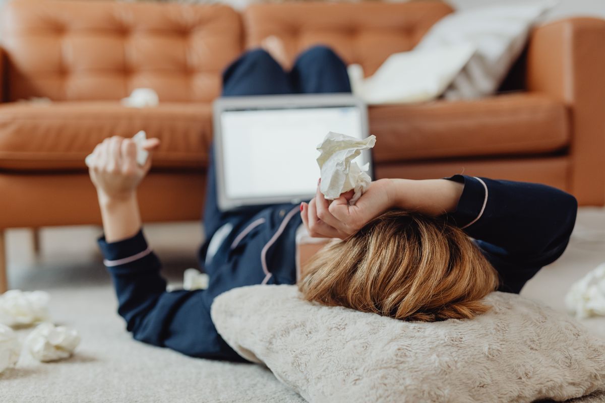 A woman lies on the floor next to a laptop, surrounded by tissues, depicting a sick day at home.