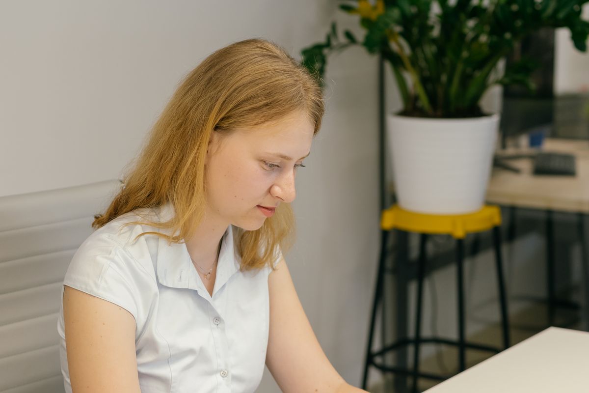 A woman in a modern office reading documents at a desk beside an indoor plant.