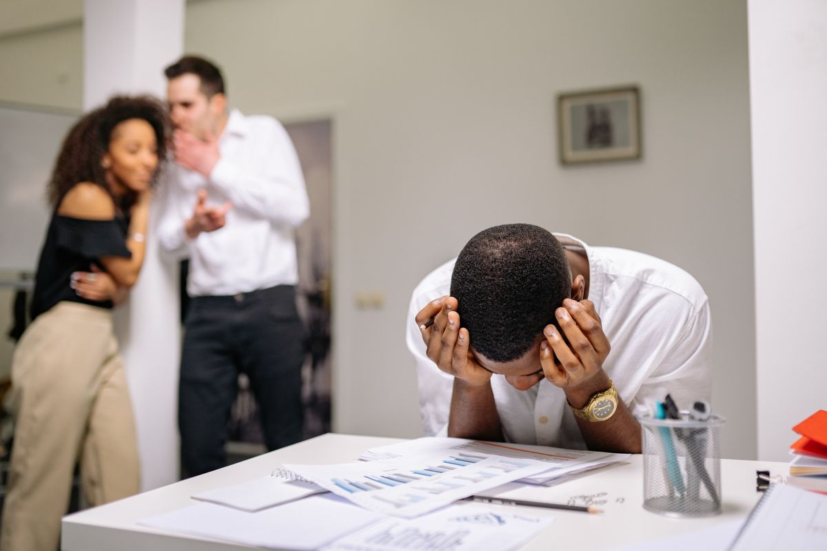 Employee with his head in his hands at a desk while colleagues whisper behind him