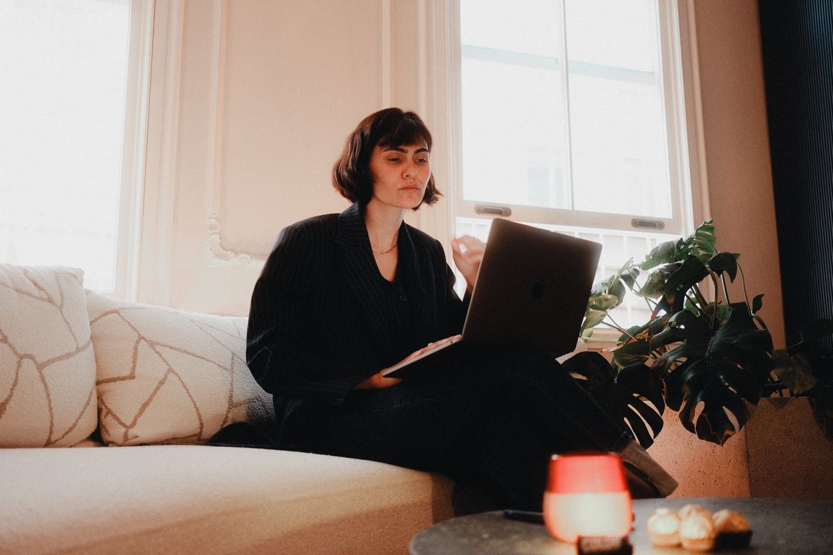 Woman in a suit using a laptop on a sofa in a bright room.