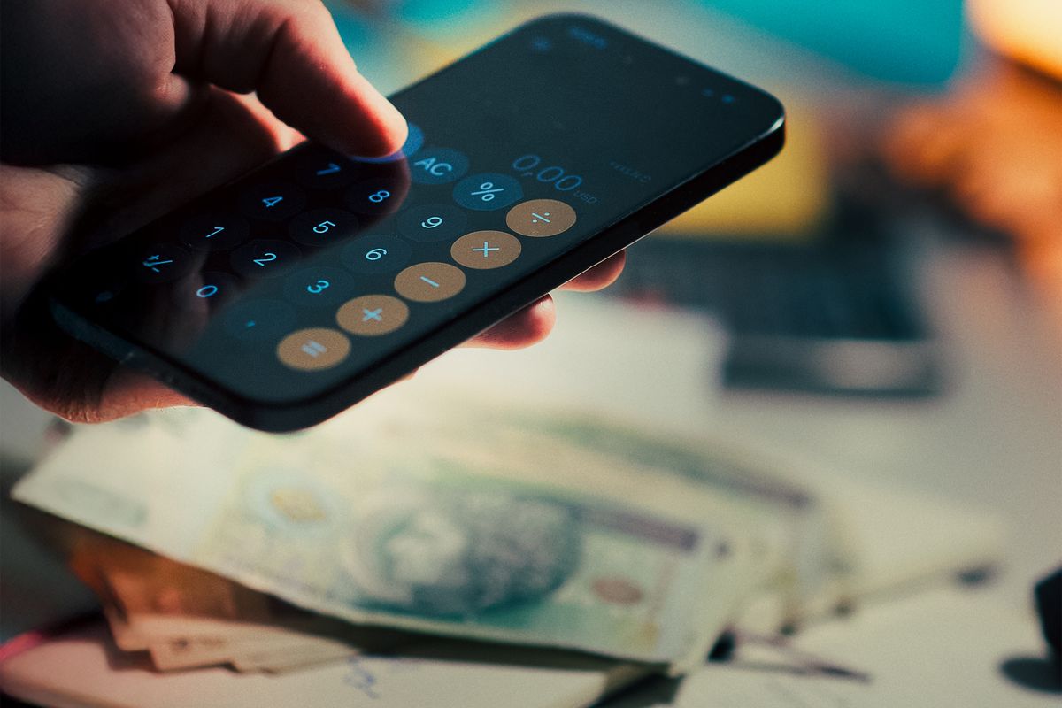A hand holds a smartphone running a calculator app, with cash and a notebook on a desk, symbolizing budgeting and finance management.