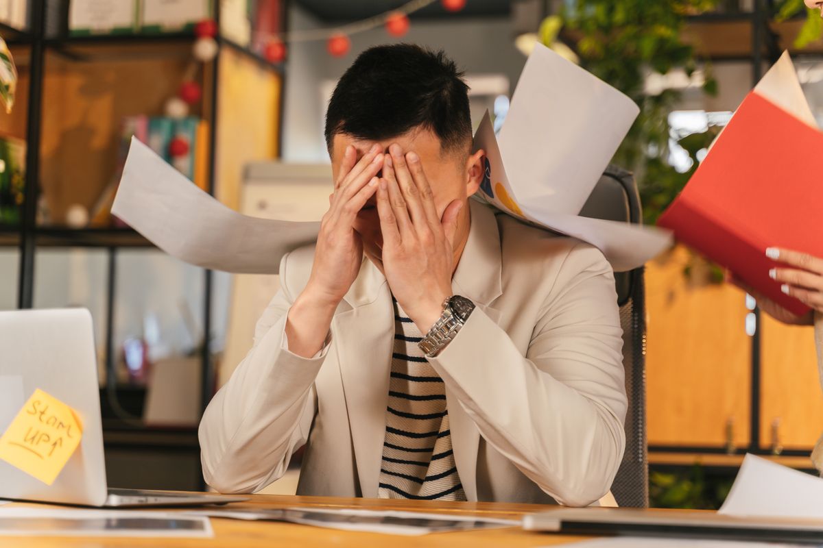 Overwhelmed employee covering his face as papers fly around his desk