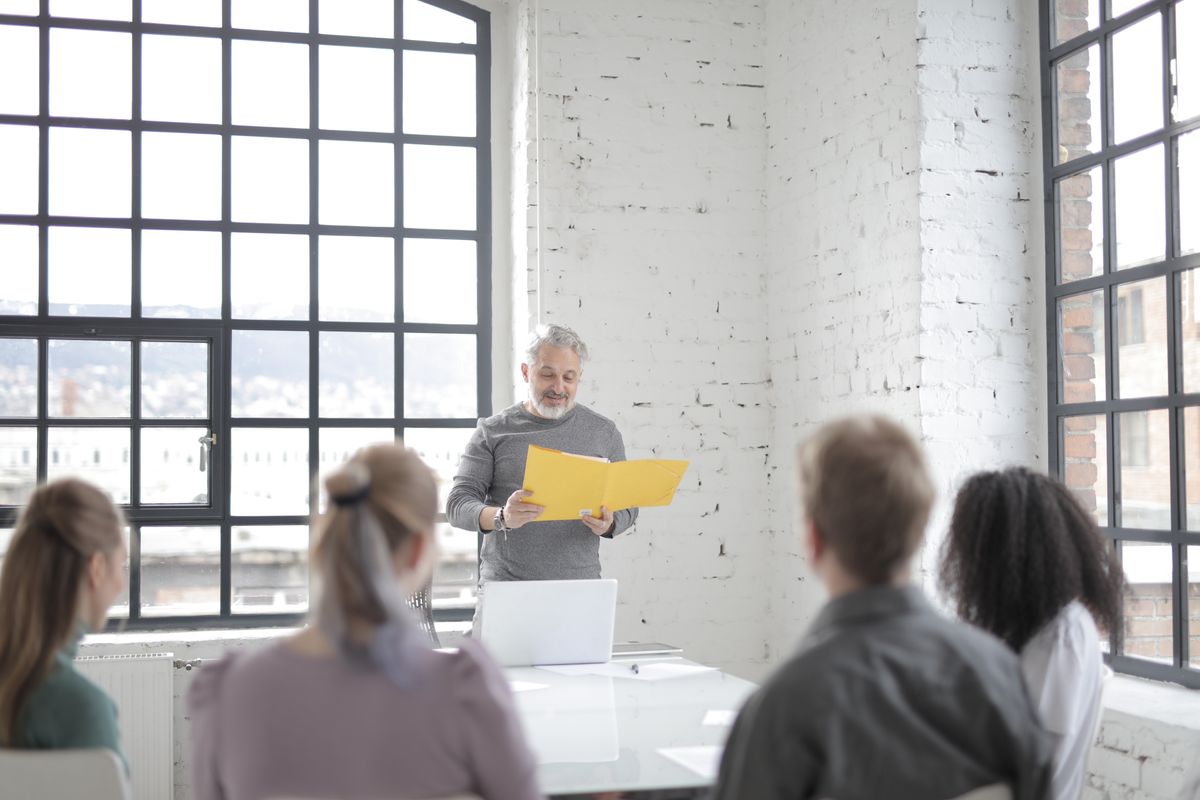 Experienced mentor presenting ideas to a group at a table with a laptop in a creative office.